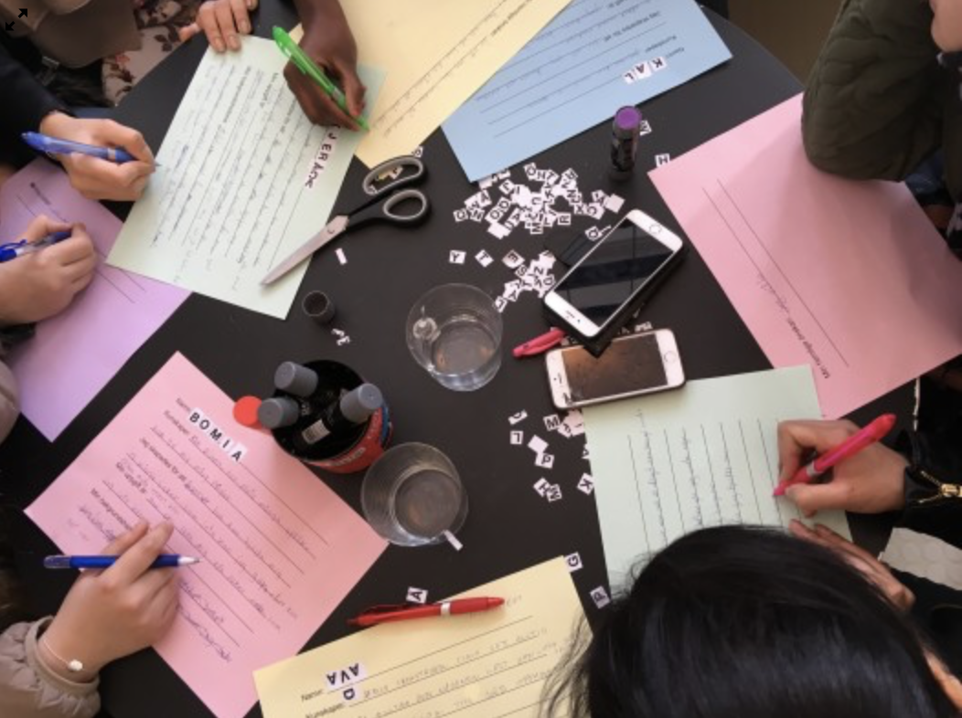 A group of people around a table full of scissors, mobile phones, markers and glues are writing on colorful papers.