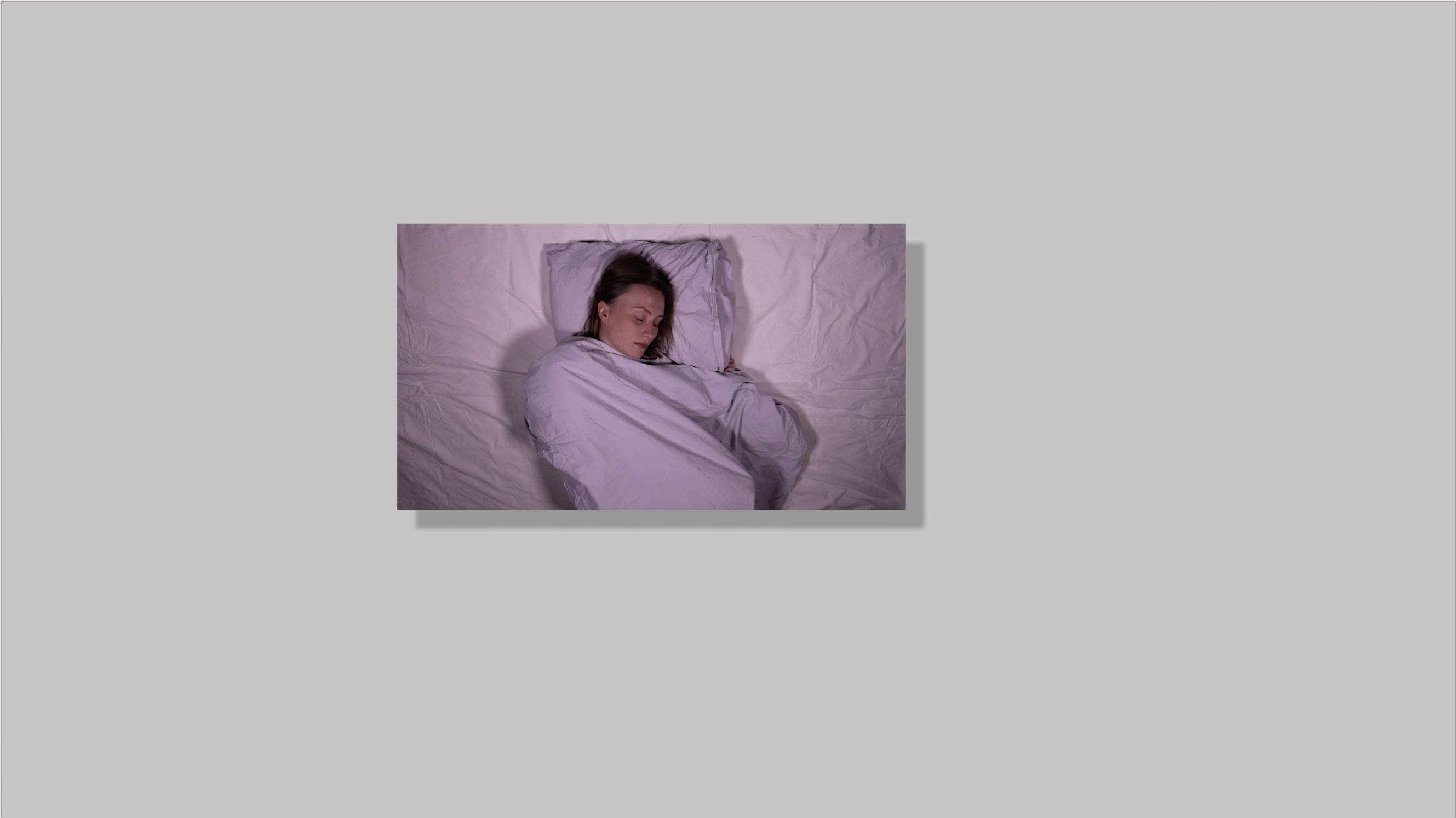 An image with a black drop shadow effect hovering on top of and in the middle of a gray background. In the image a person is sleeping in violet bed sheets. The person has brown hair, light skin and a white shirt.