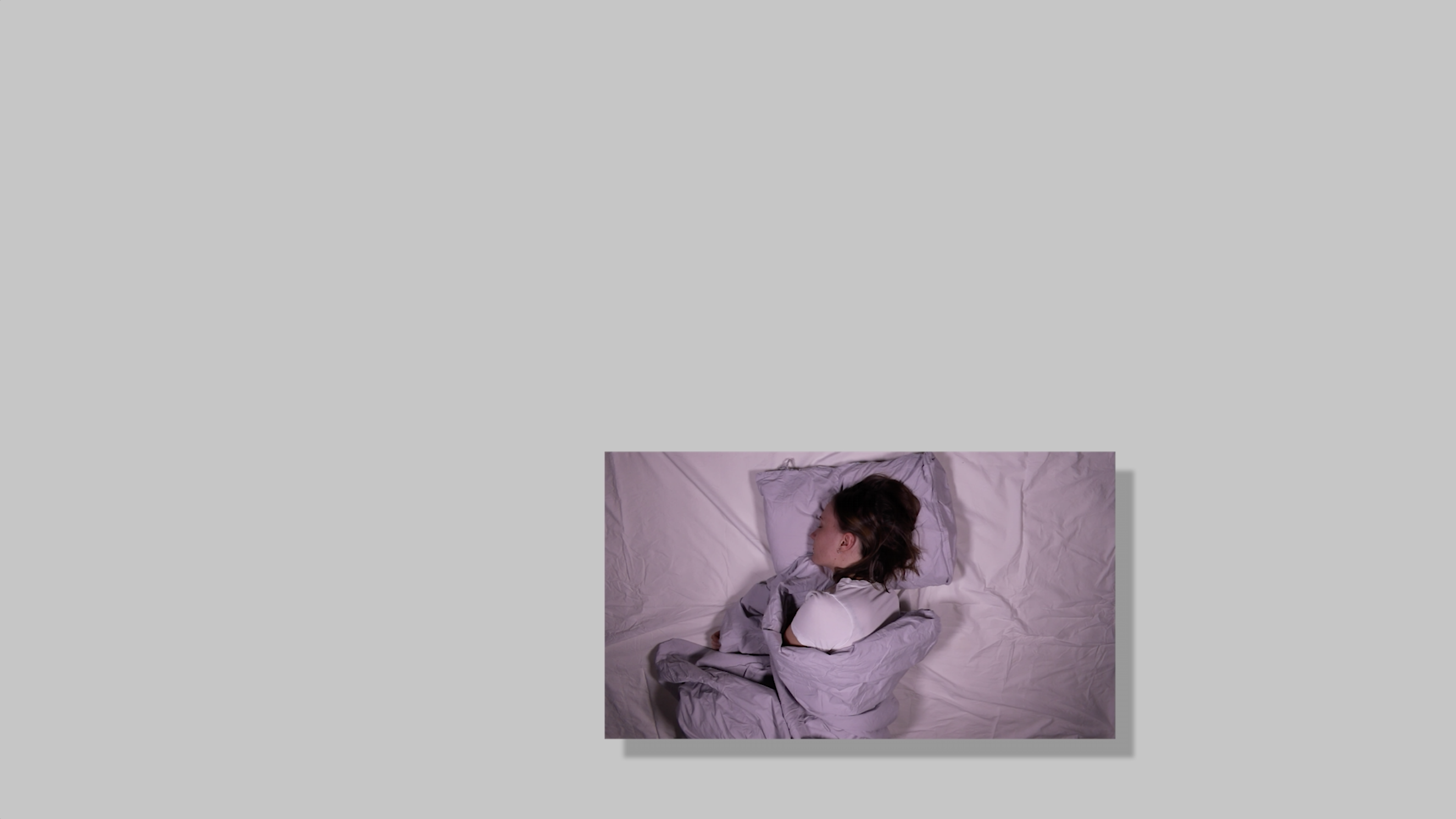 An image with a black drop shadow effect hovering on top of and at the bottom of a gray background. In the image a person is moving in violet bed sheets. The person has brown hair, light skin and a white shirt.