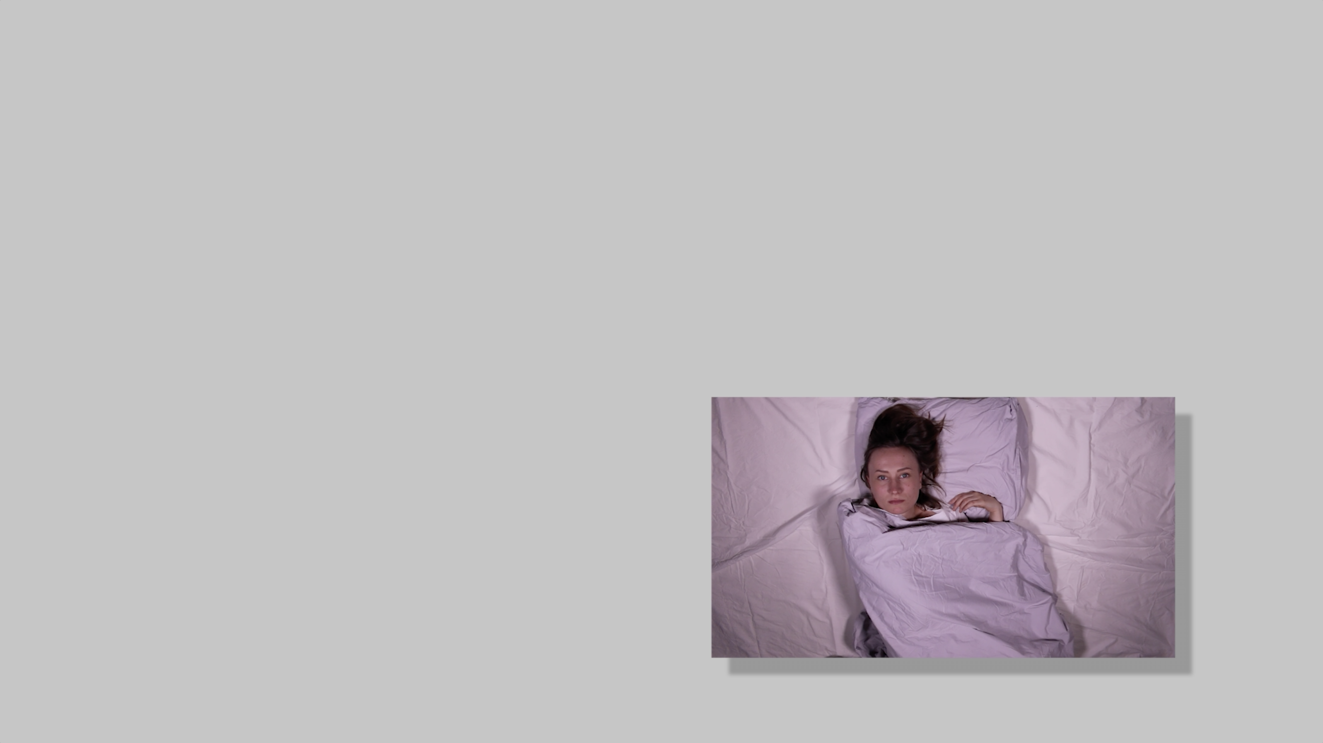 An image with a black drop shadow effect hovering on top of and at the right bottom corner of a gray background. In the image a person is laying in violet bed sheets facing the camera with their eyes open. The person has brown hair, light skin and a white shirt.